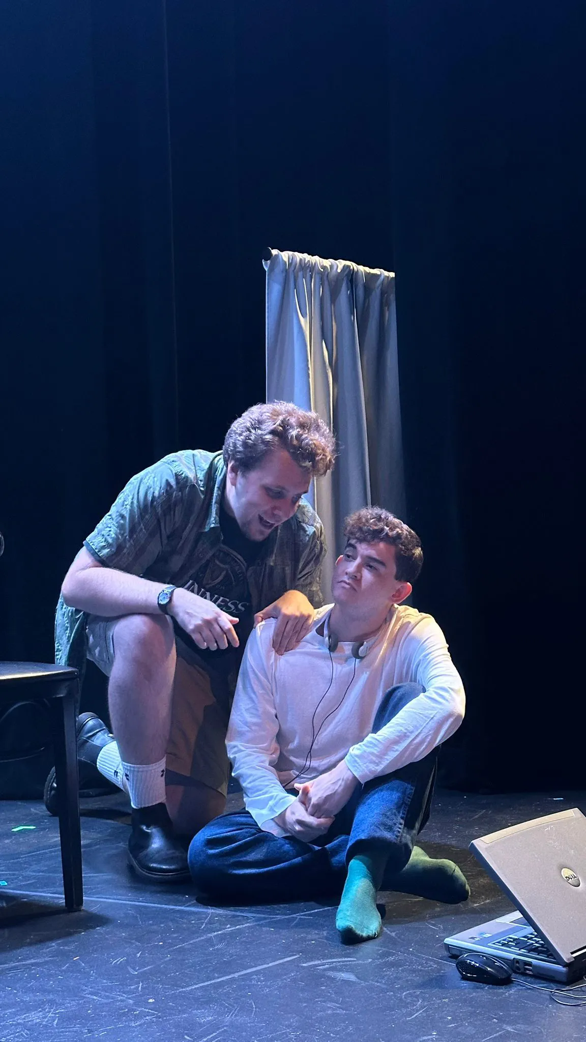 One actor crouches beside the other, who is seated on the floor of the stage.