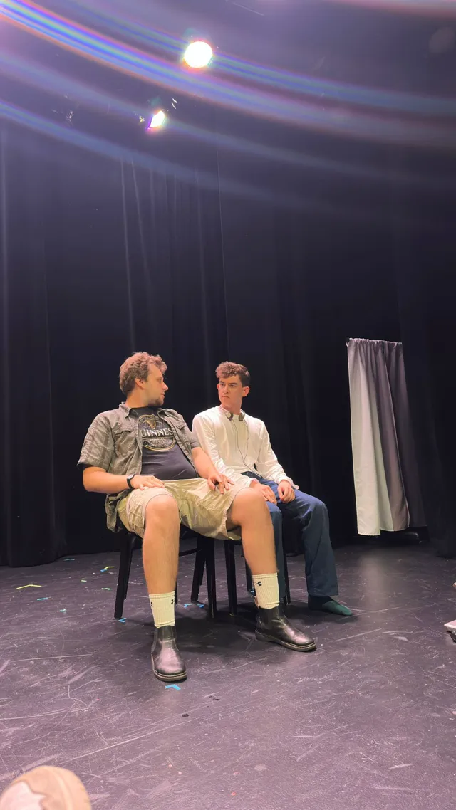 Two actors sit side by side on folding chairs on a bare stage in Beelzepup.
