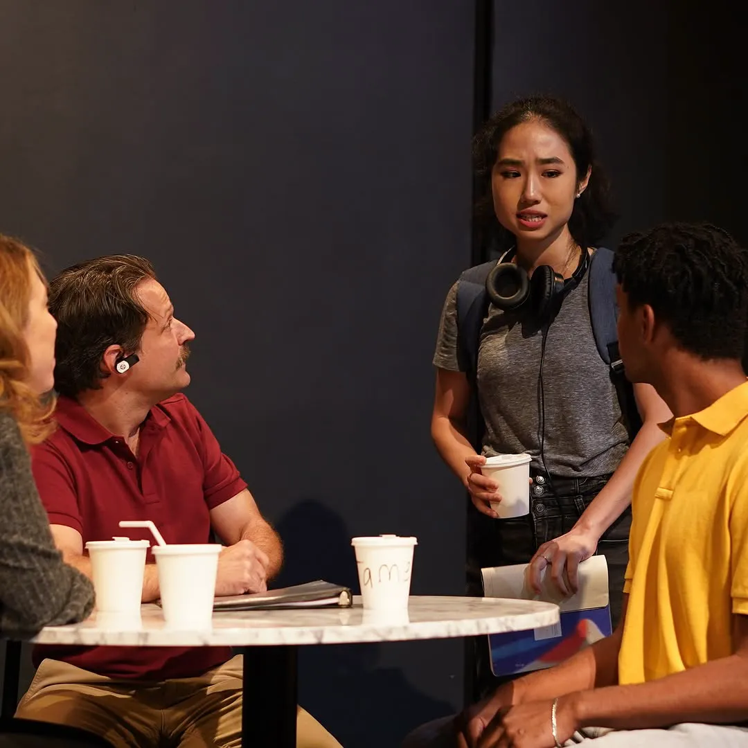 Four characters around a cafe table in the bookstore, mid-argument.