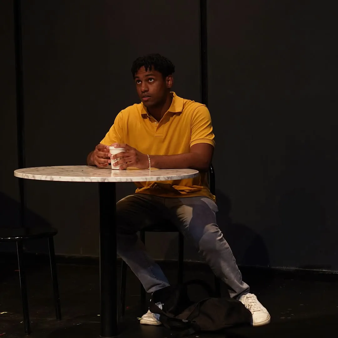 A young man in a yellow polo sits alone at the cafe table with a cup.