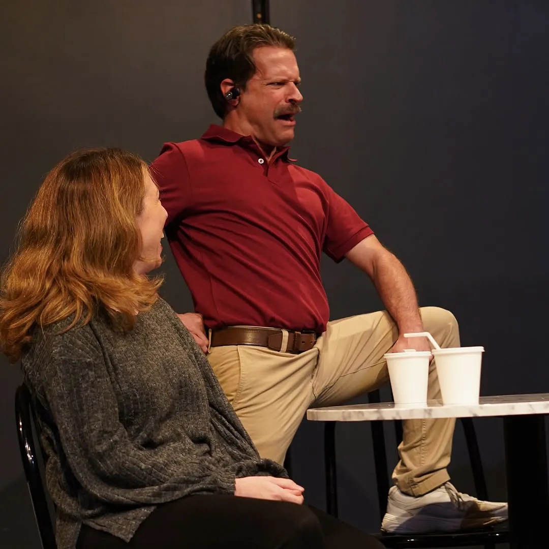 A man perches on the edge of a table beside a seated woman, both laughing.
