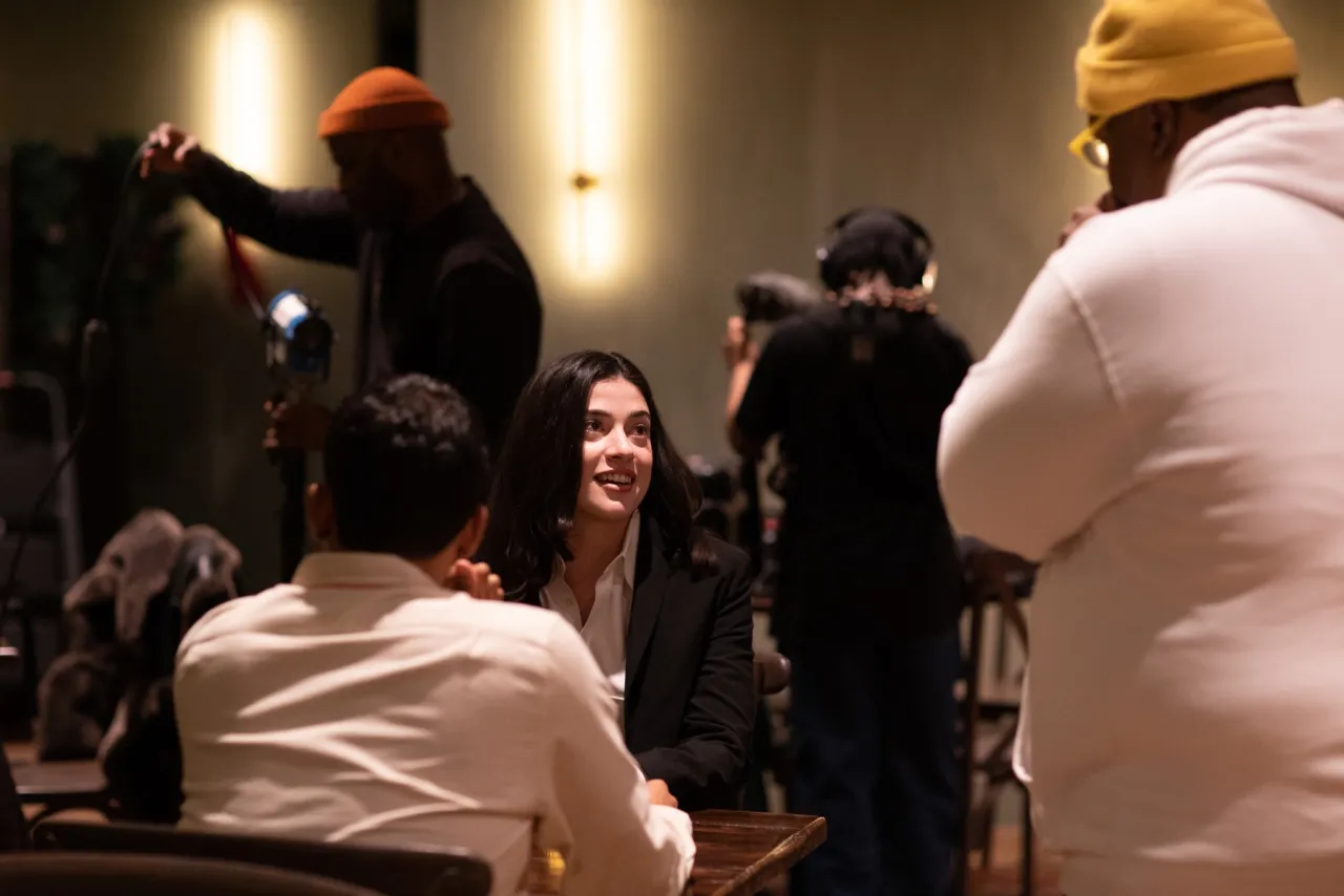 A crew films two actors seated at a table, a boom operator and camera operator working just outside the frame.
