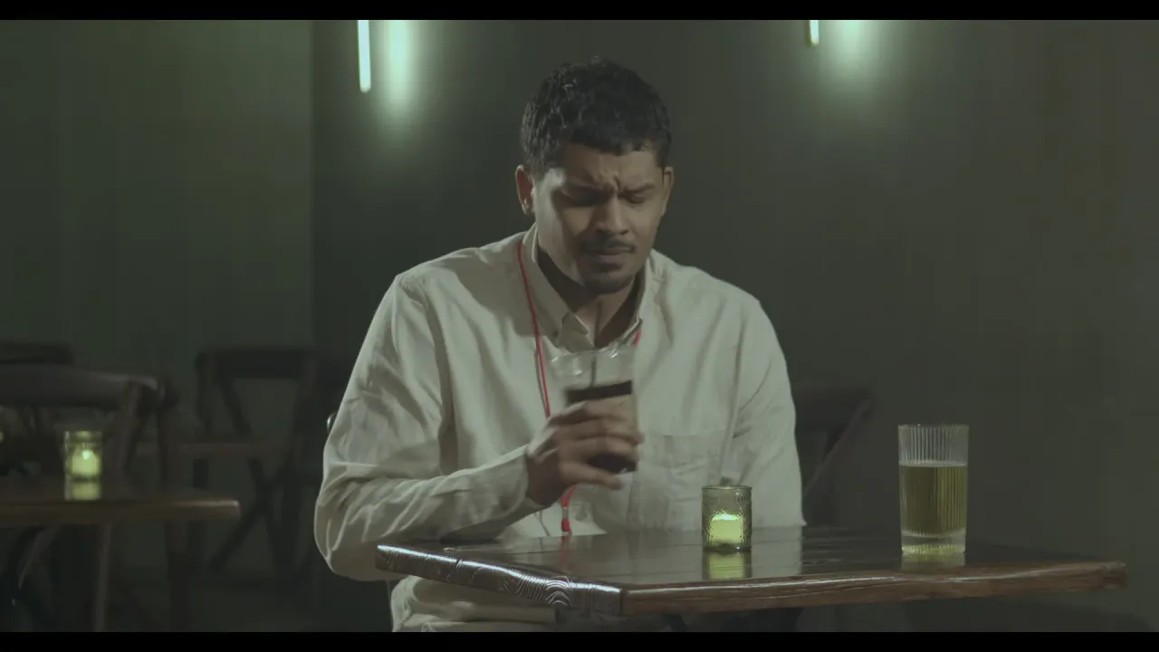 A man sits alone at a bar table under green practical lights, looking at his phone.
