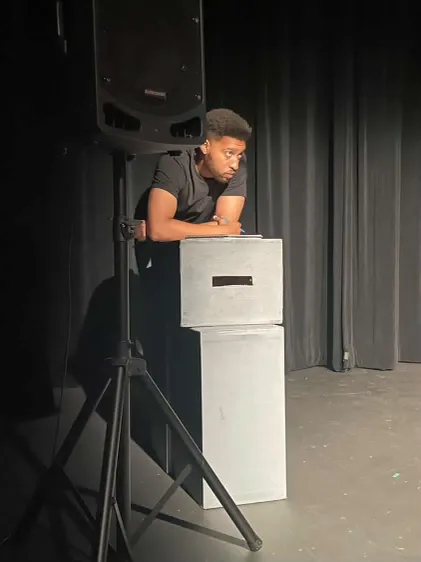 An actor leans over a white plinth on a bare stage beside a speaker on a stand.